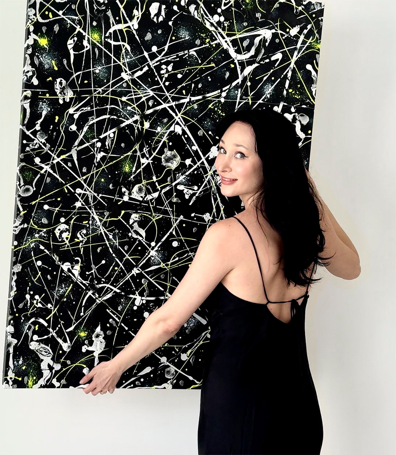 Bridget Bradley wearing black dress, holds 'Space Rock' her original abstract expressionist painting on canvas in hues of black, silver, white and neon yellow.
