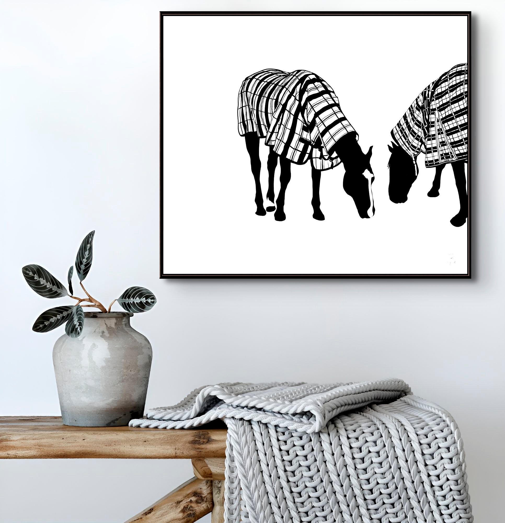 Framed artwork of two horses wearing plaid raincoats on a wall above a wooden bench with a vase and blanket. Limited edition Fine Art Print. Image © Bridget Bradley Art