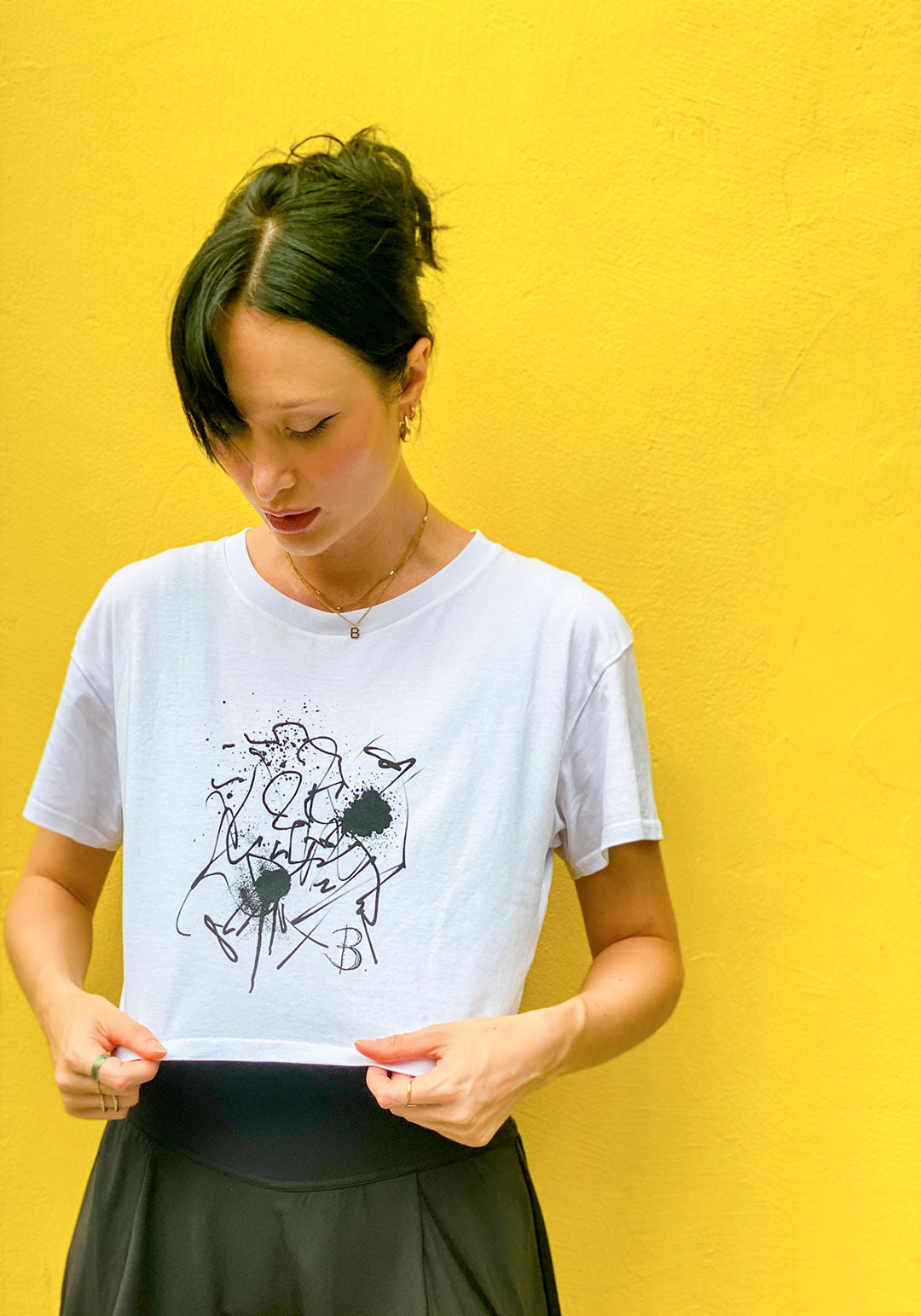 B. Contemporary Streetwear, Model Stands against yellow background wall, wears The Eva Crop Top Tee showing front., Black Design by Bridget Bradley