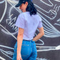 Model stands against a grey and white painted wall wearing the Smile Crop Top- Tee Showing the Back Logo from the B. Contemporary Streetwear collection