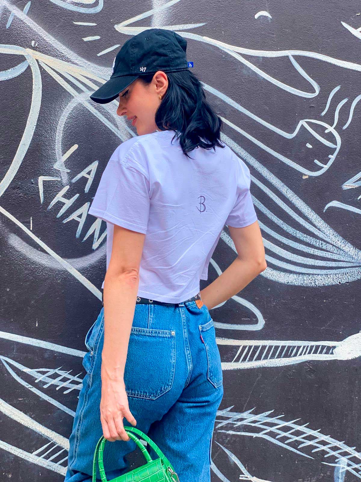 Model stands against a grey and white painted wall wearing the Smile Crop Top- Tee Showing the Back Logo from the B. Contemporary Streetwear collection