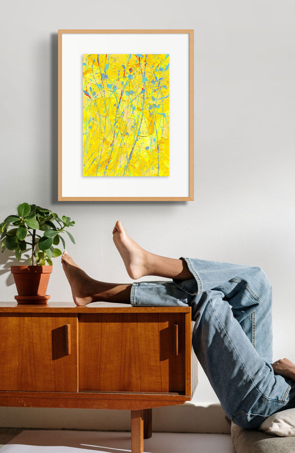 ;Sunflower' original painting, contemporary abstract art on paper by Bridget Bradley. Seen framed in oak hanging in situ above wooden console with person in jeans and feet up.